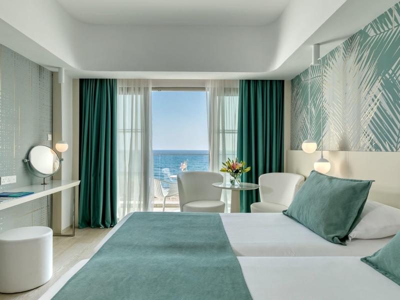 Helles Hotelzimmer mit Doppelbett, grün-weißer Dekoration und Blick auf das Meer.
