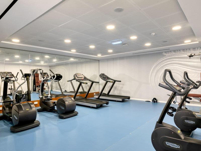 Hell beleuchteter Fitnessraum mit blauen Boden, Laufbändern, Ergometern und Kraftmaschinen.