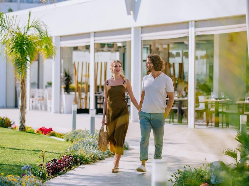 Paar hält Hände und spaziert entspannt vor modernem Haus mit großem Garten und Glasfassade.