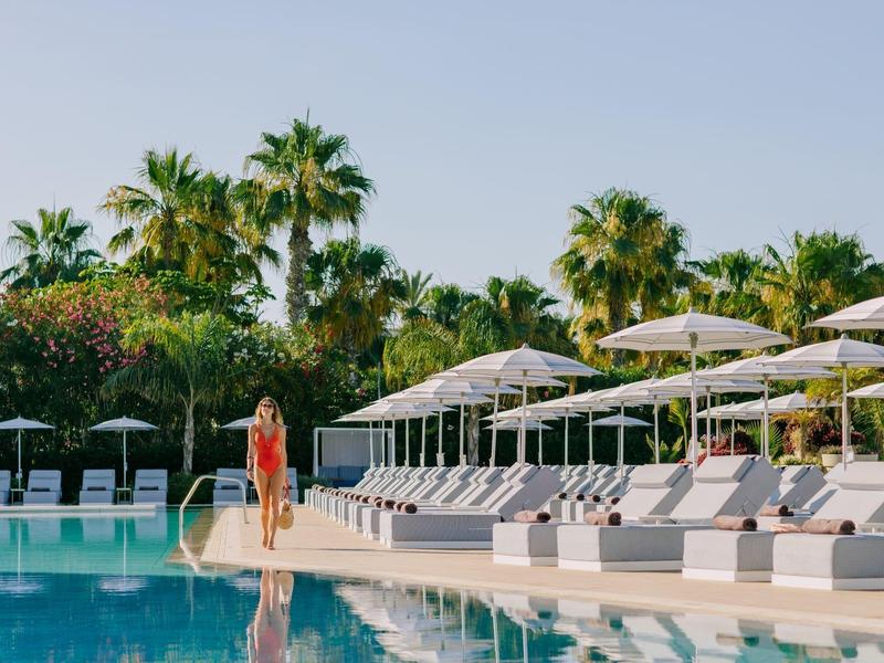 Schwimmbad mit Liegestühlen und Sonnenschirmen, Palmen im Hintergrund, Frau in rotem Kleid am Beckenrand.