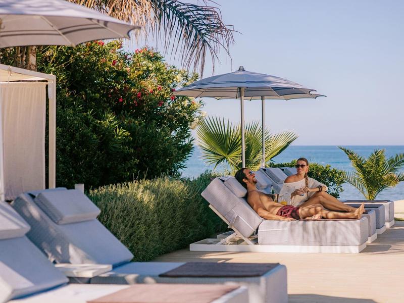 Zwei Personen entspannen auf weißen Liegen mit Sonnenschirm am Pool nahe Palmen und Meer.