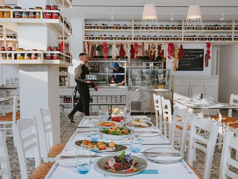 Helles Restaurant mit weißen Möbeln, langen gedeckten Tischen und Wandregalen voller Gewürzgläser.