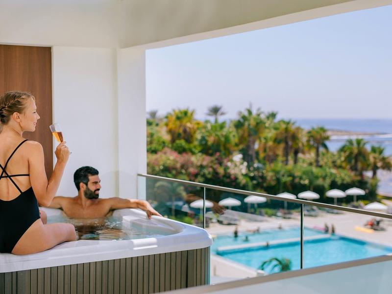 Frau und Mann entspannen im Whirlpool mit Blick auf Pool, Palmen und Meer bei klarem Himmel.
