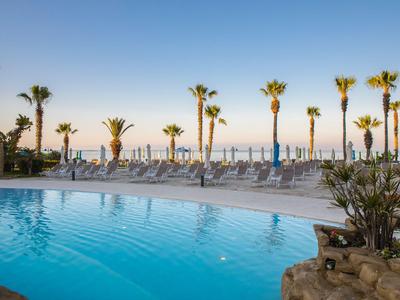 Blick auf einen Hotelpool mit Liegestühlen und Palmen unter klarem Himmel am Meer.