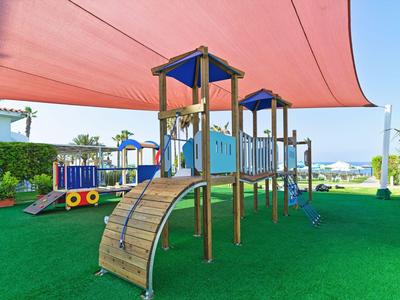 Kinderspielplatz mit Holzgerüst, Kletterrampen und Sonnensegel auf grünem Kunstrasen.