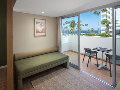 Modernes Hotelzimmer mit Sofa und Balkon mit Tisch und Stühlen, Blick auf Palmen.