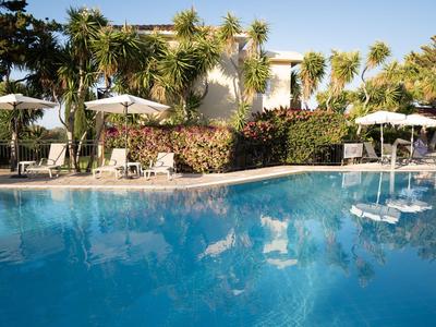Una piscina dell'hotel con acqua blu chiara, circondata da palme e lettini con ombrelloni bianchi.