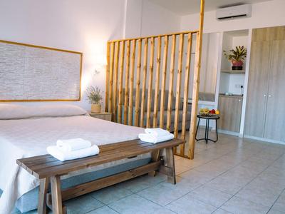 Camera d'hotel luminosa con letto matrimoniale, panca in legno, piante e doccia moderna.