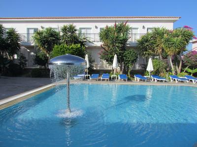 Großer blauer Pool mit Wasserspiel, umgeben von Liegestühlen, Sonnenschirmen und Gebäuden im Hintergrund.