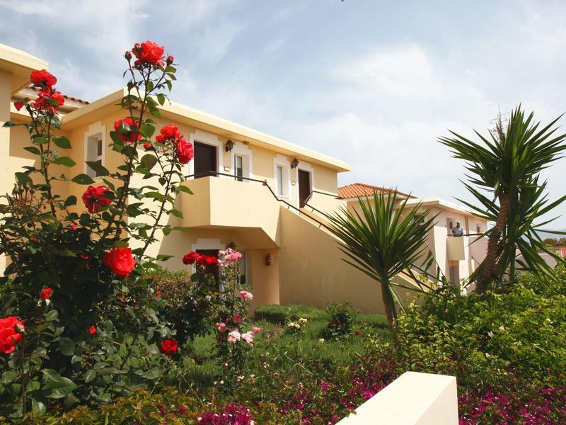 Ein beigefarbenes Gebäude mit roten Rosen und grünen Palmen im Vordergrund unter blauem Himmel.