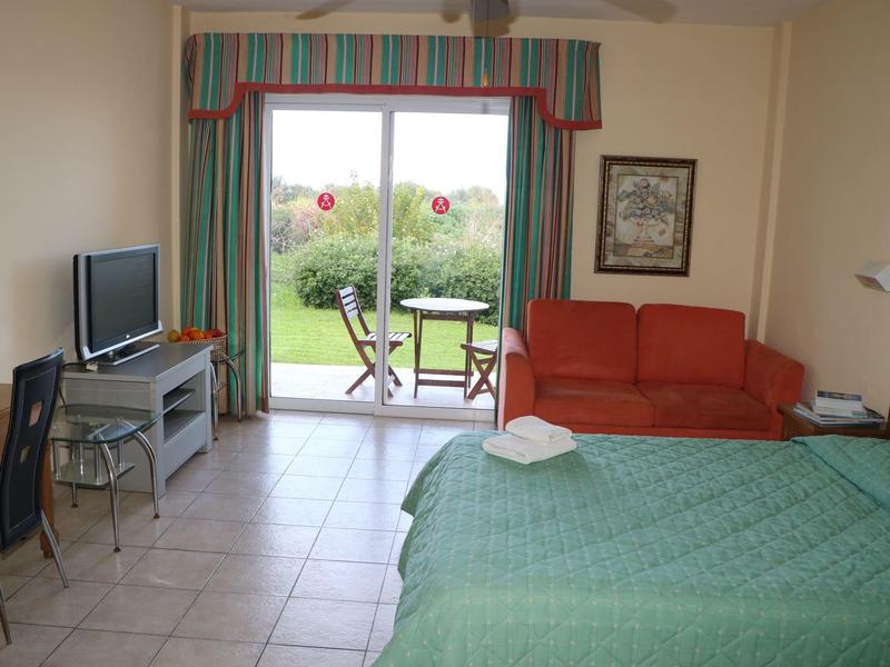 Ein helles Zimmer mit grünem Bett, rotem Sofa, TV, Tisch, Stühlen und Blick auf Garten.