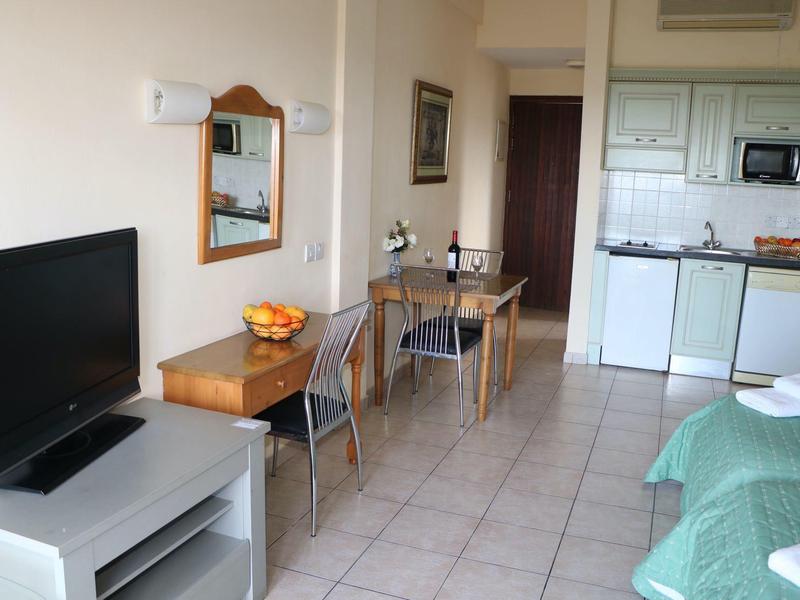 Kleines Zimmer mit Fernseher, Tisch, Stühlen, Küchenzeile und einem Bett mit grünem Bezug.