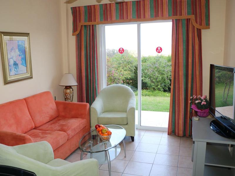 Wohnzimmer mit rotem Sofa, zwei grünen Sesseln, Glastisch, Fernseher und großer Fensterfront mit Blick ins Grüne.