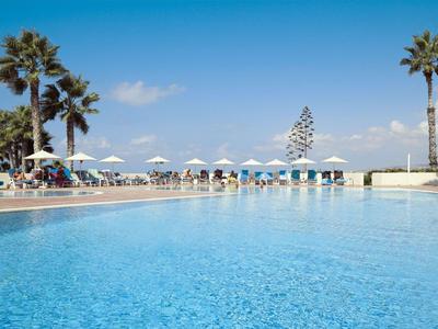 Groot blauw zwembad met palmbomen en witte parasols onder een heldere lucht.