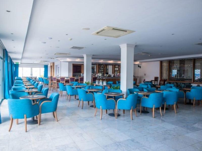 Großer heller Gastraum mit blauen Sesseln, weißen Tischen und großen Fenstern mit blauen Vorhängen.