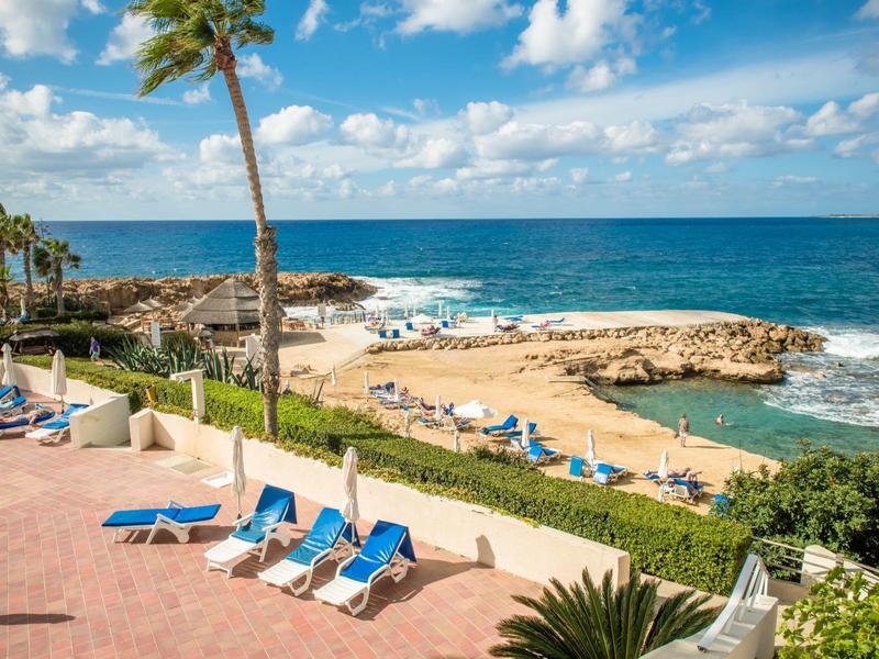 Meerblick mit Felsen, blauem Himmel, Palmen und blauen Liegestühlen auf einer Terrasse am Strand.