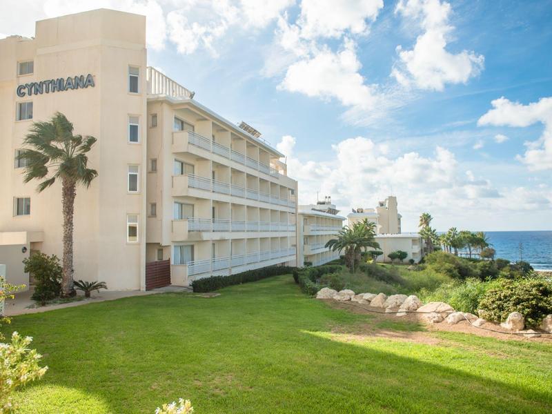 Ein mehrstöckiges Hotelgebäude mit Palmen vor, grünem Rasen und Meer im Hintergrund unter blauem Himmel.