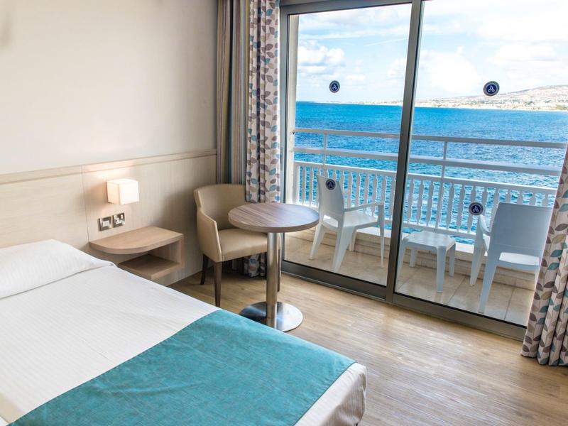 Hell eingerichtetes Hotelzimmer mit Einzelbett, kleinem Tisch, zwei Stühlen und Balkon mit Meerblick.