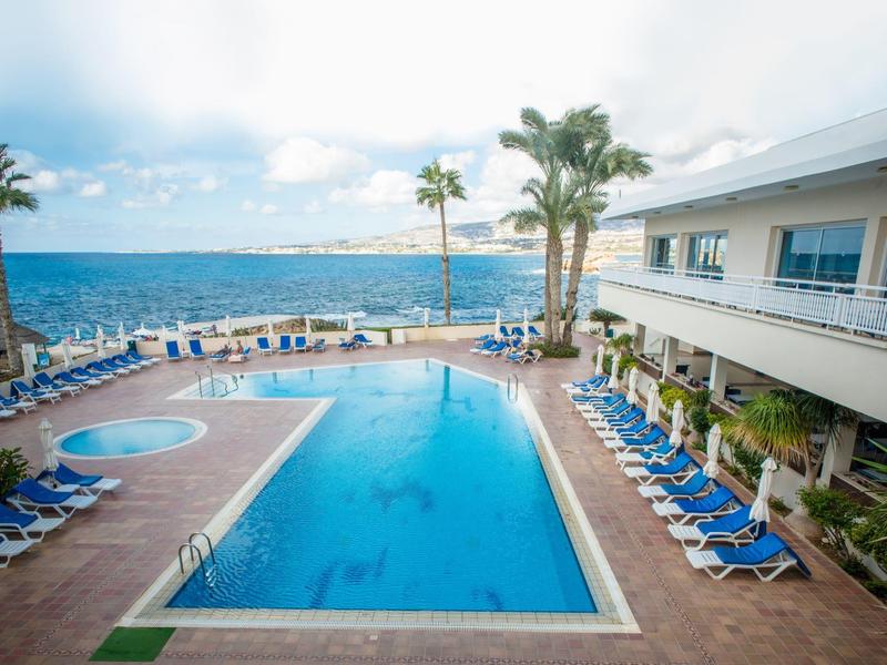Großer Pool mit blauen Liegestühlen, Palmen und Blick aufs Meer an einem sonnigen Tag.