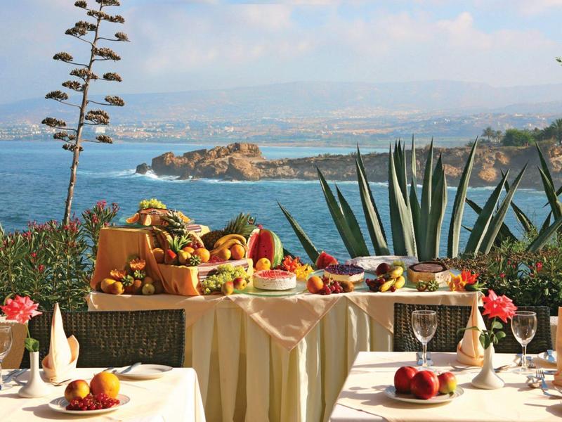 Gedeckter Tisch mit Früchten und Getränken vor blauem Meer und grüner Küstenlandschaft.