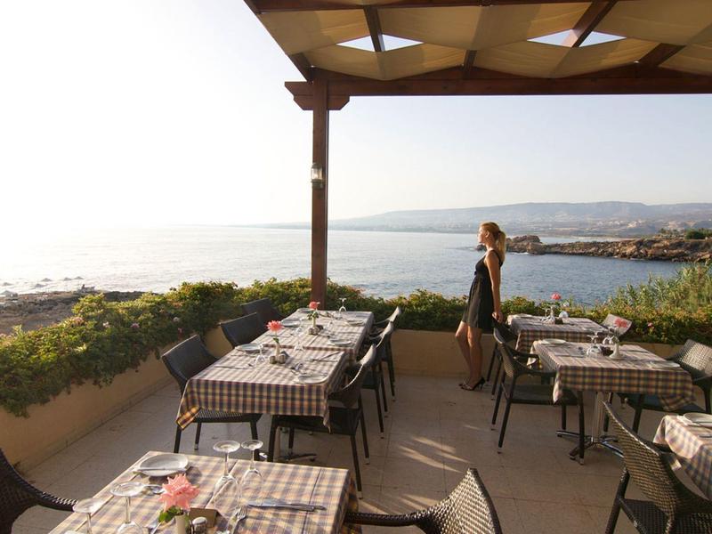 Terrassenrestaurant mit karierten Tischdecken, Küstenblick und einer stehenden Frau im Sonnenlicht.