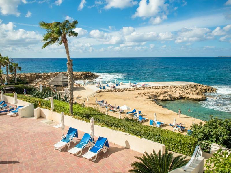 Strand mit Liegestühlen, Palmen und blauem Meer unter einem bewölkten Himmel.