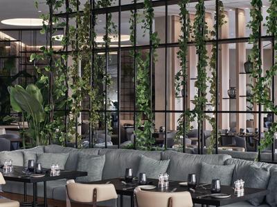 Modernes Café mit grünen Kletterpflanzen, grauen Sofas und schwarzen Tischen in hellem Innenraum.