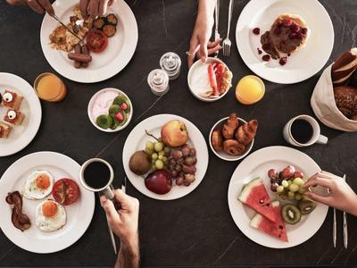 Verschiedene Teller mit Früchten, Gebäck und Getränken auf dunklem Tisch, mehrere Menschen essen.