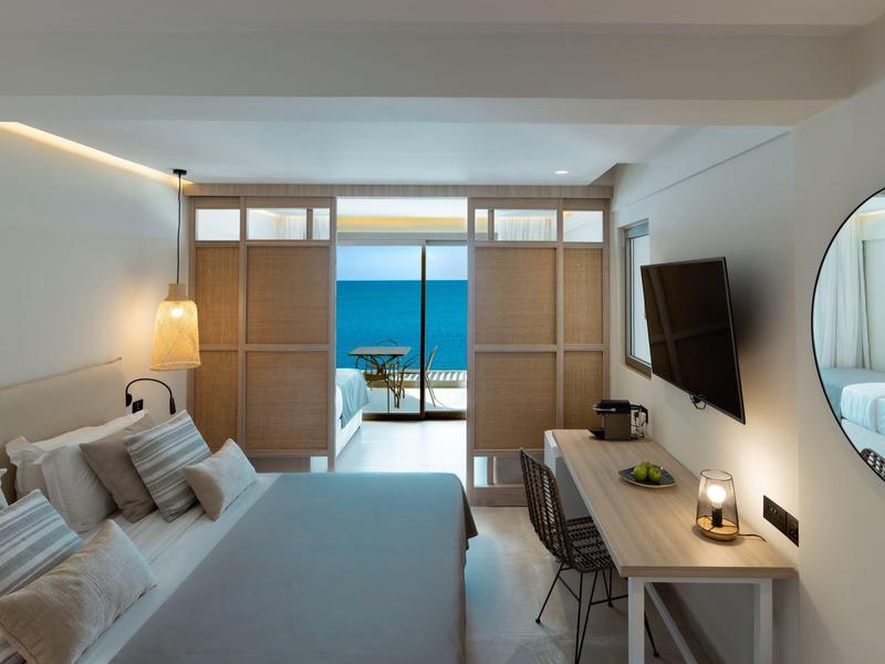 Hell eingerichtetes Schlafzimmer mit großem Fenster und Meerblick, modernem Schreibtisch und TV.