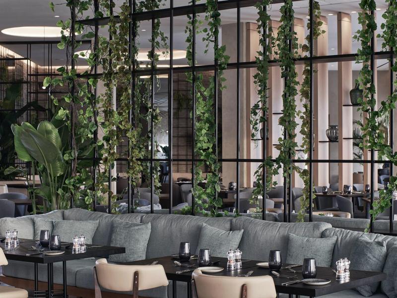 Modernes Café mit grünen Kletterpflanzen, grauen Sofas und schwarzen Tischen in hellem Innenraum.