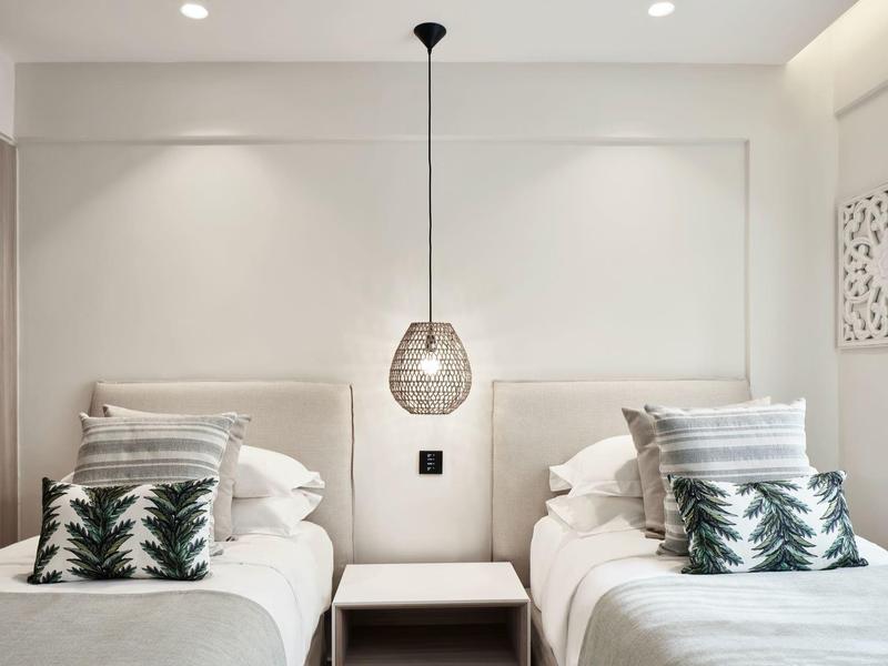 Minimalistisches Schlafzimmer mit zwei Einzelbetten, weißen Wänden, grünen Kissen und Hängeleuchte in der Mitte.