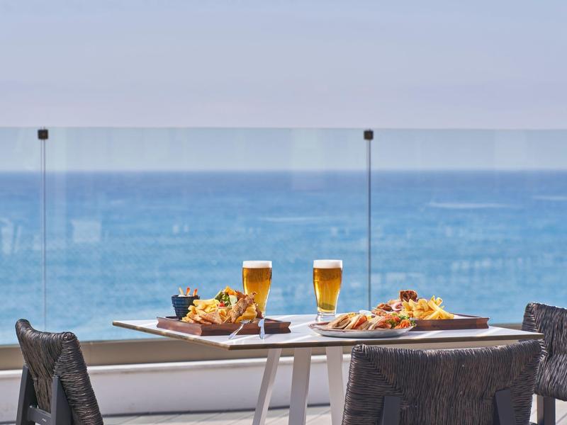 Balkontisch mit Essen und Bier vor Glasgeländer, dahinter blauer Ozean und heller Himmel.