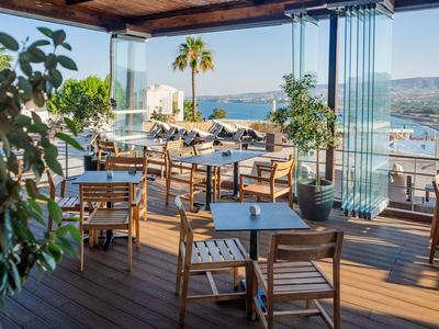 Offene Terrasse mit Holzmöbeln und Meerblick, von Pflanzen und gläsernem Schutz umgeben.