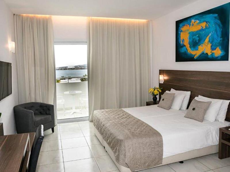 Hell eingerichtetes Hotelzimmer mit Doppelbett, dunkelgrauem Sessel und Balkon mit Blick auf Wasser.