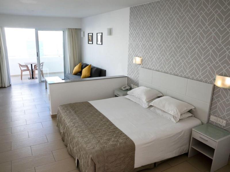 Helles Schlafzimmer mit großem Bett, Nachttisch, gemusterter Wand und Blick auf Balkon mit Meer.