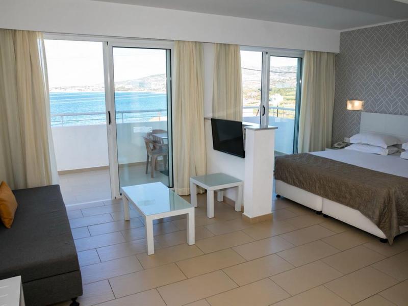 Helles Hotelzimmer mit Doppelbett, Sofa, TV, Glas-Couchtisch und Balkon mit Meerblick.