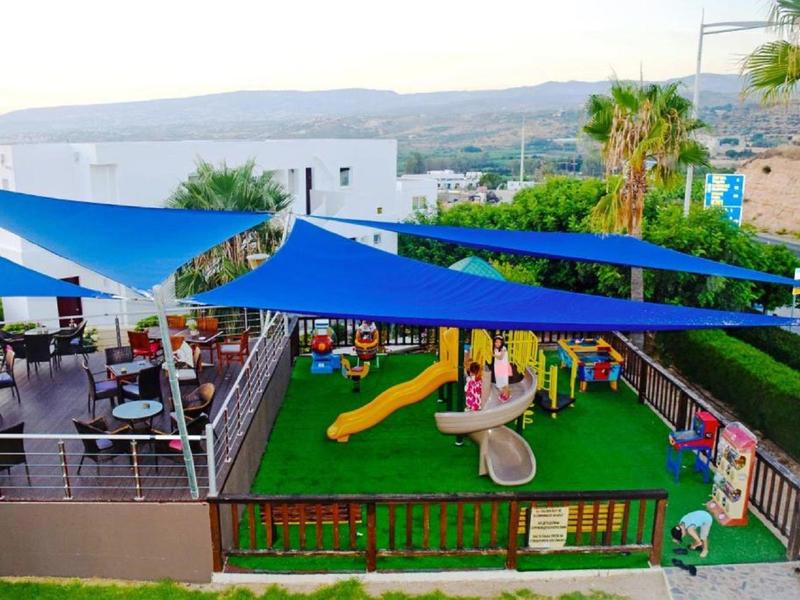 Außenbereich mit blauem Sonnensegel, Spielplatz mit Rutsche und Sitzbereich mit Tischen und Stühlen.