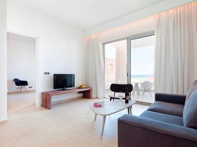 Heller moderner Wohnbereich mit Sofa, TV, Minibar und großem Fenster mit Vorhängen.