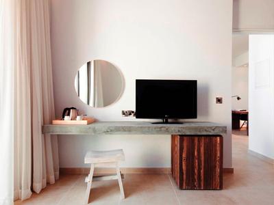 Minimalistischer Raum mit Flachbildfernseher, rundem Spiegel und Hocker vor einer Fensterfront.