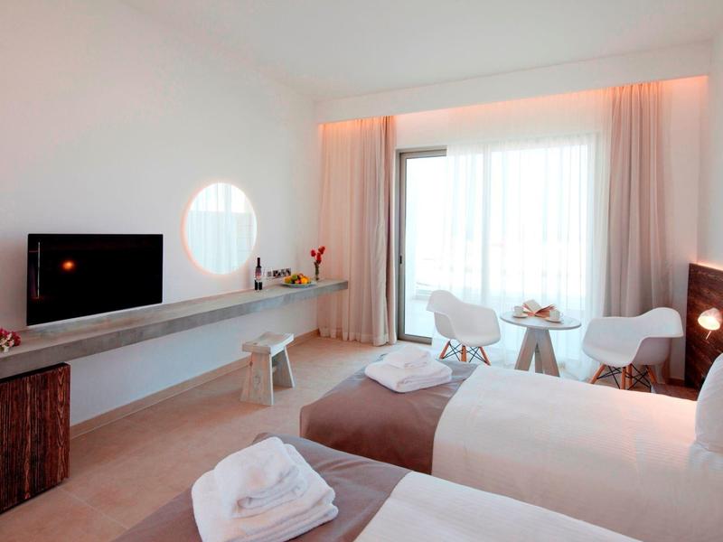 Helles Hotelzimmer mit zwei Betten, TV, Schreibtisch und Balkon mit Vorhängen.