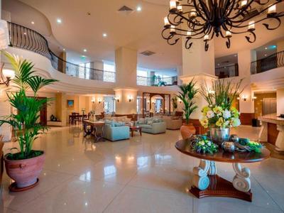 Prostorná hotelová lobby s vysokým stropem, rostlinami a dekorativním lustrem.