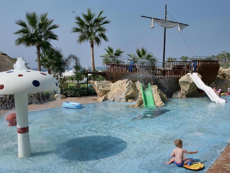 Kinder plantschen in einem flachen Pool mit Wasserrutsche und Palmen im Hintergrund.