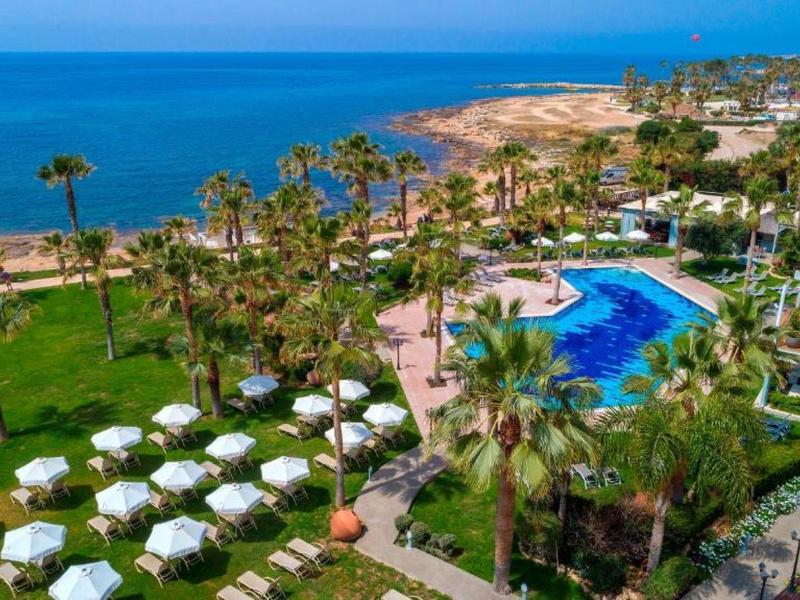 Hotelanlage mit Pool, grünen Palmen und weißen Sonnenschirmen an einer Küste am Meer.