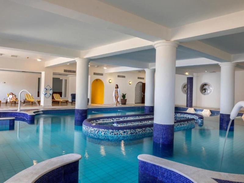 Innenraum mit großem runden Schwimmbecken, weißen Säulen und blau-weißen Fliesen.