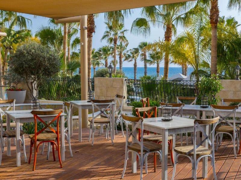 Leere Terrasse mit weißen Tischen und Stühlen, umgeben von Palmen und Blick aufs blaue Meer.