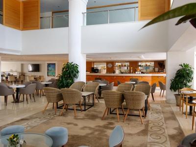 Hell beleuchteter Hotel-Loungebereich mit Tischen, Sesseln und Pflanzen.