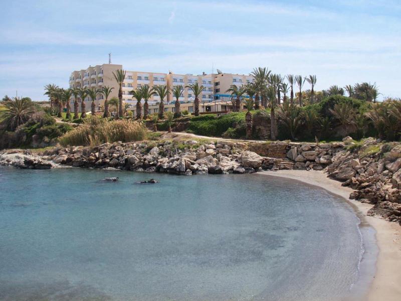 Eine ruhige Bucht mit klarem Wasser und Sandstrand, umgeben von Felsen und Palmen vor Gebäuden.