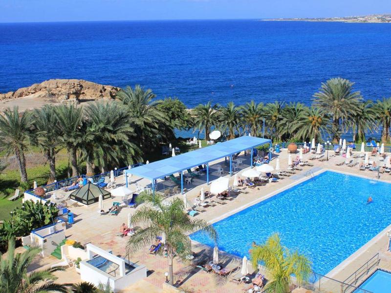 Großer rechteckiger Pool mit Liegestühlen, Palmen und blauem Meer im Hintergrund.
