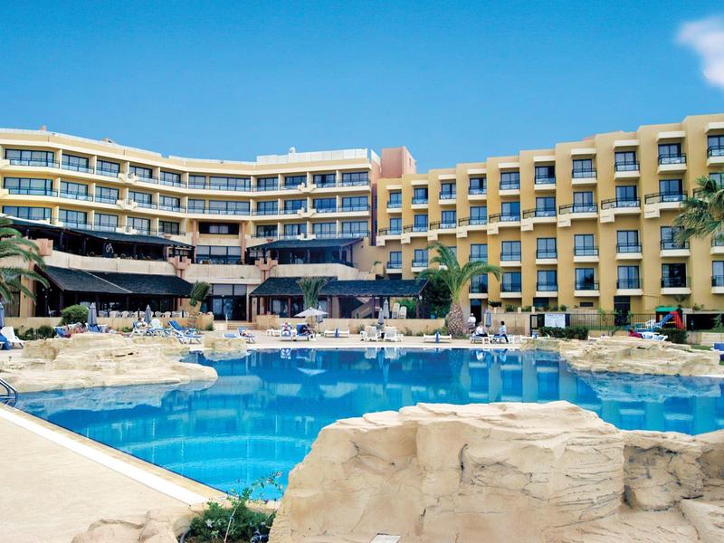 Großes Hotel mit klarblauem Pool, Liegestühlen und Palmen unter strahlend blauem Himmel.