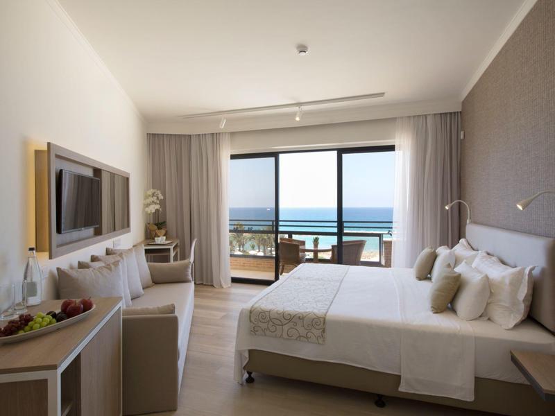 Helles Hotelzimmer mit großem Bett, TV, Sitzbereich und Balkon mit Meerblick.